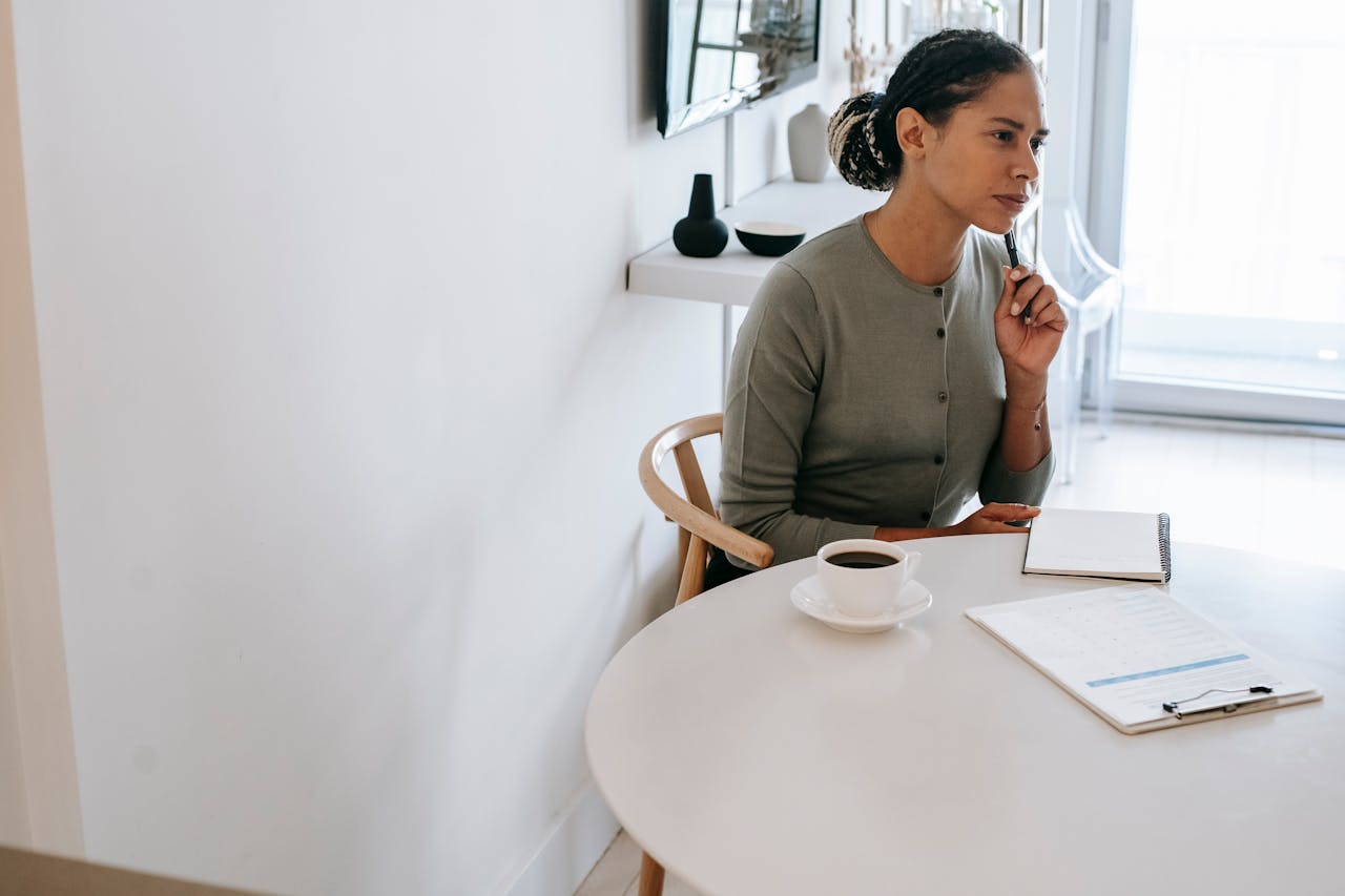 Concentrated ethnic female HR interviewer or psychologist in formal clothes sitting at round table with pen and notepad while looking away in contemplation